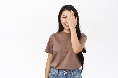 Portrait of young asian woman cover one side of face, looking with one eye, half covered with palm, smiling, checking vision at optician store, white backgroundの写真素材