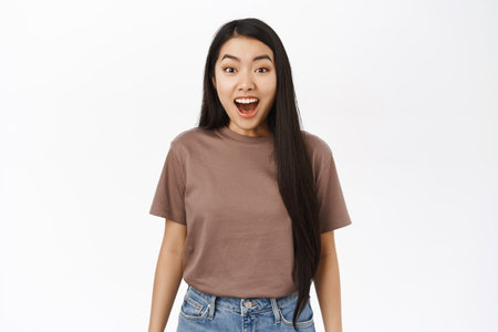 Surprised asian woman gasping, looking amazed at camera, impressed face, standing over white backgroundの写真素材