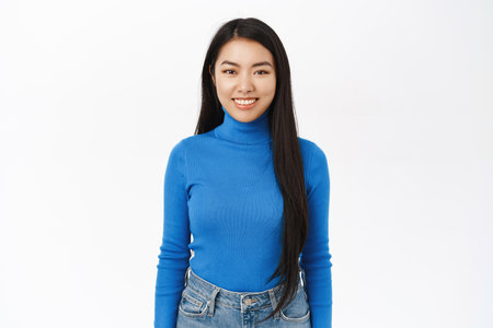 Stylish asian smiling woman, wearing blue long sleeve, looking happy and confident, standing over white backgroundの写真素材