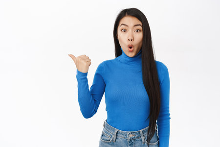 Awesome promo offer. Surprised asian woman pointing finger left, gasping and saying wow, impressed by smth, standing over white backgroundの写真素材