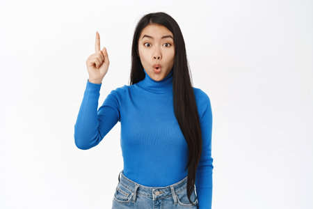 Enthusiastic asian female model pointing finger up, shows advertisement, pitching an idea, eureka gesture, stands over white backgroundの写真素材