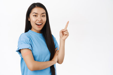 Enthusiastic asian girl got vaccinated, has band aid patch on shoulder after covid vaccine, smiling and pointing at copy space banner, showing coronavirus info announcementの写真素材