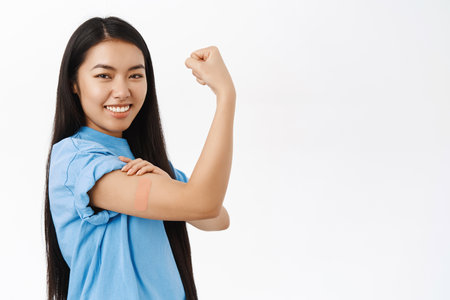 Smiling asian woman with band aid on shoulder, did covid-19 vaccination, flexing biceps, feeling strong after vaccine, white backgroundの写真素材