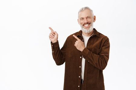 Enthusiastic old man, senior mature person pointing fingers left, smiling and laughing, showing advertisement, standing against white backgroundの写真素材