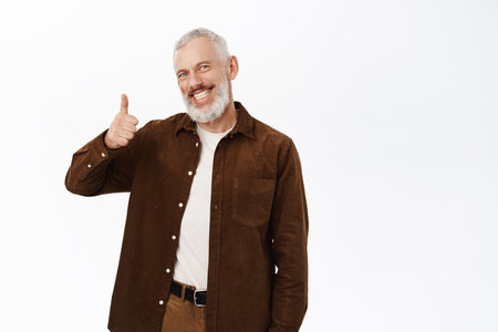 Smiling mature man, senior male model with grey beard, showing thumbs up, recommending smth good, praise, compliment, standing over white backgroundの写真素材