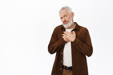 Image of senior guy, old man being touched, holding hands on chest grateful, looking flattered and pleased, standing over white backgroundの写真素材