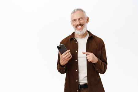 Handsome mature man with white beard, pointing finger at smartphone, showing mobile phone app, standing over white backgroundの写真素材