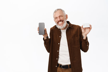 Senior man showing smartphone screen and credit card, smiling pleased, standing in casual clothes over white backgroundの写真素材