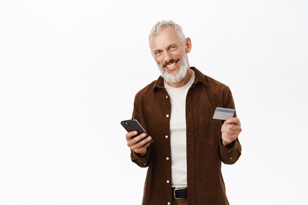 Portrait of handsome senior man, caucasian old guy using smartphone and holding credit card, shopping online, paying with mobile phone, white backgroundの写真素材