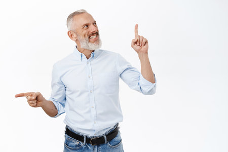 Portrait of senior business man pointing up and left, showing two promos, standing in blue shirt and jeans over white backgroundの写真素材