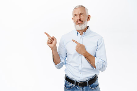 Portrait of caucasian senior guy, businessman pointing fingers left and looking at copy space advertisement, standing over white backgroundの写真素材