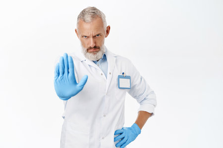 Image of senior doctor, male physician showing stop sign, prohibition gesture, stopping you, standing over white backgroundの写真素材