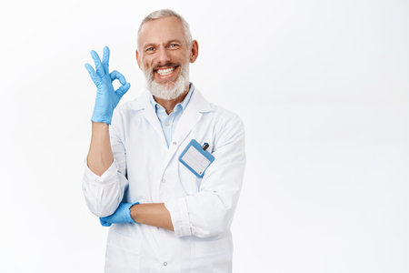 Smiling senior doctor, medical worker showing okay sign, recommending clinic, standing over white backgroundの写真素材