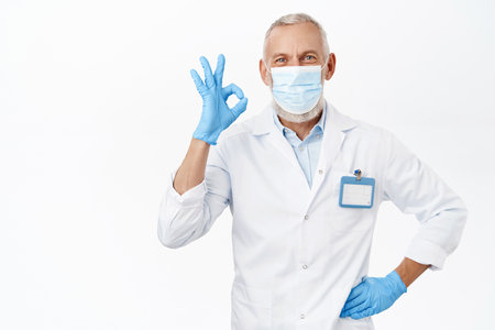 Coronavirus and health concept. Handsome senior man, doctor or dentist showing okay sign, approve smth, recommending, white backgroundの写真素材
