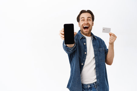 Online shopping and mobile banking concept. Smiling happy guy shows smartphone screen and credit card, stands over white backgroundの写真素材