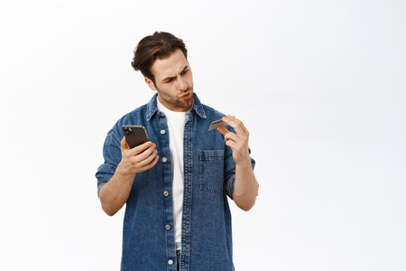 Portrait of man looking confused at his credit card, has problem paying on mobile phone app, standing over white backgroundの写真素材