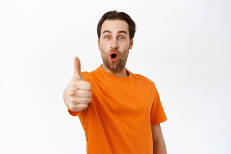 Man with impressed face shows thumbs up, recommends smth awesome, stands over white backgroundの写真素材