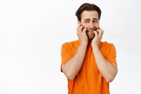 Scared adult man biting his fingernails, looking anxious, standing over white backgroundの写真素材