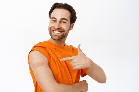 Covid vaccination campaign. Smiling healthy adult man shows his shoulder with band aid from coronavirus vaccine shot, standing in orange tshirt over white backgroundの写真素材