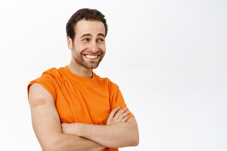 Handsome healthy guy smiles, shows shoulder with band aid after covid 19 vaccination shot, vaccine injection, stands over white backgroundの写真素材