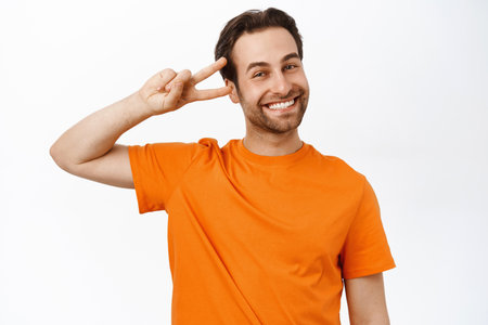 Portrait of smiling brunette man shows peace, v-sign and looks happy, expresses joy, stands in orange tshirt over white backgroundの写真素材