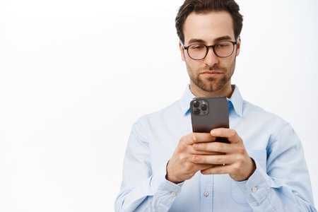 Serious male entrepreneur looking at his mobile phone, reading on smartphone, using application, standing in glasses against white backgroundの写真素材