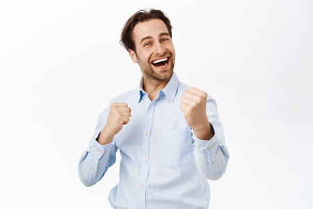 Handsome triumphing man fist pump, celebrating victory or achievement, smiling pleased, standing over white backgroundの写真素材