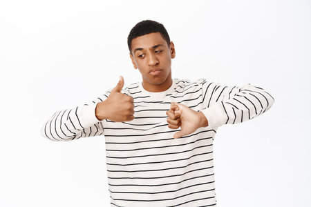 Portrait of smiling african american man shows thumbs up and thumbs down, like or dislike, negative and positive, making decision, standing over white backgroundの写真素材