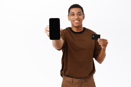 African american man shows his mobile phone screen and credit card, demonstrates online banking application, shopping website, standing over white backgroundの写真素材