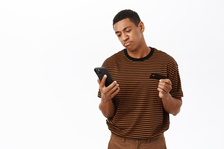 African american man looking disappointed, using mobile phone and credit card, standing in casual clothesの写真素材