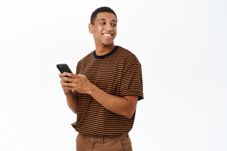 Mobile technology concept. Smiling african american man using his smartphone, holding phone and looking happy, texting in application, white backgroundの写真素材