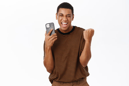 Excited Black man winning on mobile phone, holding smartphone and celebrating success, standing over white backgroundの写真素材