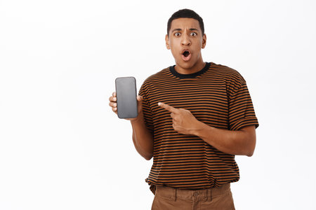 Shocked and disappointed African american guy pointing at his mobile phone, showing on smartphone screen, white backgroundの写真素材