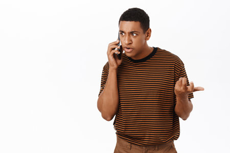 African american man arguing on mobile phone, calling person and looking confused, standing over white backgroundの写真素材