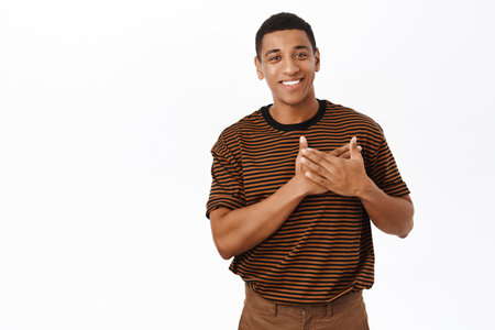 Image of smiling african american guy holds hands on heart, looking with care and appreciation, stands over white backgroundの写真素材