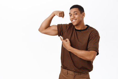 Sport and gym concept. Strong African american man flex biceps, shows muscles, looking confident, standing over white backgroundの写真素材