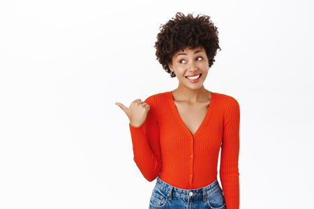 Beautiful smiling african american girl points finger left, this way gesture, shows logo or banner beside her, stands over white backgroundの写真素材