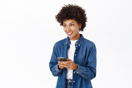 Smiling african american woman using mobile phone, holding telephone and looking happy, standing over white backgroundの写真素材