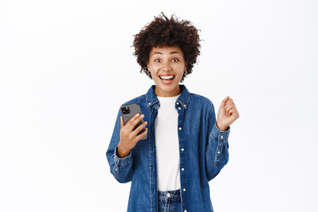 Happy beautiful young woman jumps from excitement, using mobile phone and looking joyful, winning on smartphone app, standing over white backgroundの写真素材