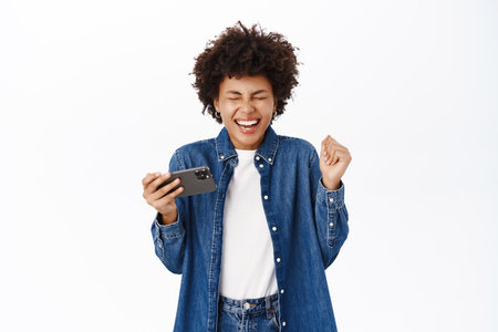 Happy beautiful young woman jumps from excitement, using mobile phone and looking joyful, winning on smartphone app, standing over white backgroundの写真素材