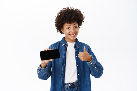 Portrait of happy young smiling woman, showing her mobile phone screen, online offer on smartphone, making thumbs up in approval, white backgroundの写真素材