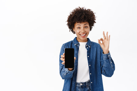 Portrait of happy young smiling woman, showing her mobile phone screen, online offer on smartphone, making okay gesture in approval, white backgroundの写真素材