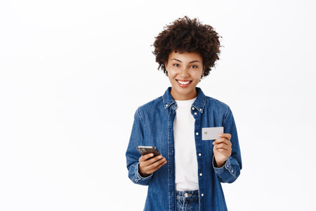 Smiling young woman looks happy while using her credit card and mobile phone. Girl with smartphone orders smth online, shops in application, white backgroundの写真素材