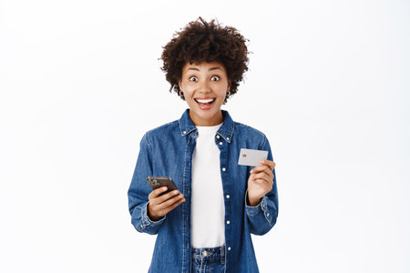 Smiling young woman looks happy while using her credit card and mobile phone. Girl with smartphone orders smth online, shops in application, white backgroundの写真素材