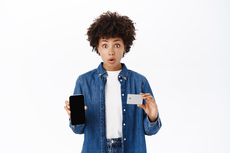 Online shopping and mobile banking concept. Happy young woman shows her phone screen and credit card with broad smile, white backgroundの写真素材