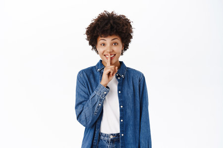 Portrait of Black woman shows hush, shhh shush gesture, taboo, asks to keep quiet, gossiping, stands over white backgroundの写真素材