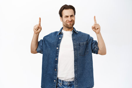Skeptical adult man, 30 years old, points up, grimaces from dislike, shows disapproval, demonstrates smth bad upwards, stands over white backgroundの写真素材