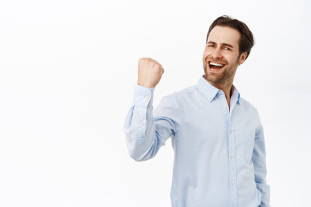 Handsome triumphing man fist pump, celebrating victory or achievement, smiling pleased, standing over white backgroundの写真素材