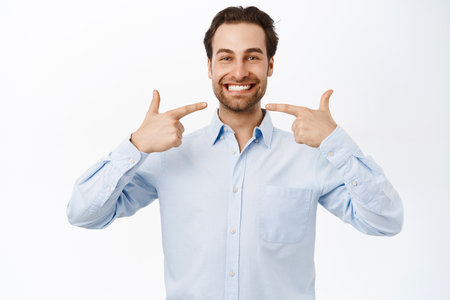 Portrait of smiling handsome businessman pointing at his white perfect smile, standing over white backgroundの写真素材