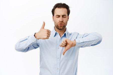 Good or bad. Male entrepreneur shows thumbs up and thumbs down, like and dislike gesture, negative or positive feedback, stands over white backgroundの写真素材